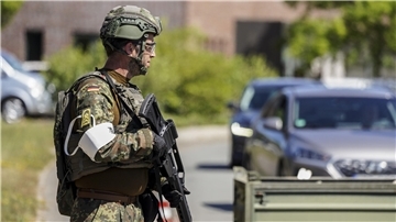 Ein Bundeswehrsoldat in Tarnuniform und Schutzhelm steht mit Gewehr an einer Straße. Im Hintergrund warten Autos vor einer Kontrollstelle.