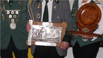 Detailaufnahme von drei Personen in Schützenuniformen. Ihre Uniformen sind mit Abzeichen und Orden geschmückt. Sie halten eine Holzplakette mit eingearbeiteter Metallgravur und eine hölzerne Ehrentafel.