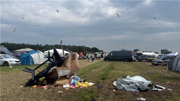 Sofas und Plastikmöbel auf einem Haufen auf einer Wiese. Drumherum Zelte und Autos. Am Himmel kreisen Möwen.