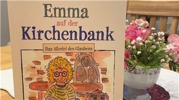 Das Foto zeigt im Vordergrund ein Buch. Dahinter ist eine kleine Vase mit Blumen zu sehen, die auf einem Tisch steht.