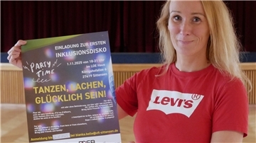 Eine blonde Frau in einem roten T-Shirt hält ein Plakat in der Hand.