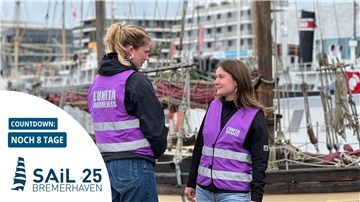 Zwei Frauen in violetten Westen stehen vor einem Schiff. Im Hintergrund Binnenhafen mit größeren Häusern.
