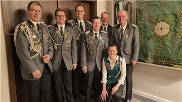 Sieben Mitglieder eines Schützenvereins posieren in festlicher Uniform vor einer Wand mit einer großen grünen Vereinsfahne. Die Männer tragen graue Jacken mit zahlreichen Orden und Abzeichen, eine Frau in der vorderen Reihe trägt eine grüne Weste mit Auszeichnungen.