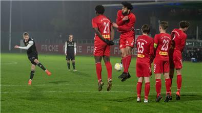Fünf Männer in roten Trikots stehen mit dem Rücken zur Kamera und ein Mann schießt einen Ball zwischen die Beine.