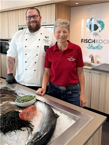 Ein Mann und eine Frau stehen vor einem Tisch, auf dem sich unter anderem ein Fisch befindet. Florian Zerbst - Fischsommelier aus dem Fischkochstudio - und Karin Steffens vom Landfrauenverein Bremervörde: Im Fischkochstudio feierte die neue „Klimakochshow“ Premiere. Fisch wird dort als genussvolle und zugleich klimafreundliche Alternative vorgestellt.