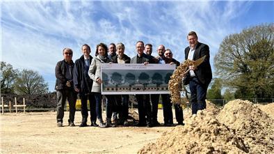 Neue Kita in Wehdel: Der erste Spatenstich ist gemacht Menschen stehen auf einem Sandfeld, drei von ihnen halten ein Plakat. Der Bürgermeister schleudert Sand von einem Spaten.