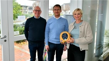 Zwei Männer und eine Frau sind auf dem Bild zu sehen. Die Frau hält einen Defibrillator in ihren Händen. 