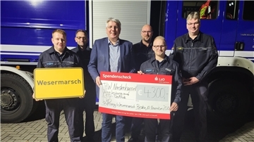 Sechs Männer lachen in die Kamera. Zwei Männer halten einen Spenden-Scheck in der Hand. 