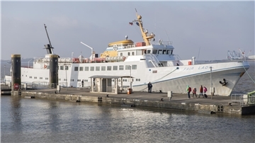 Ein Schiff liegt an der Kaje. Ein paar Fahrgäste sind mit Rucksäcken und Koffer im Anmarsch. 
