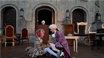 Auf einer Theaterbühne drei Personen in barocken Kostümen. Eine Frau und ein Mann im Vordergrund albern herum, im Hintergrund ein Mann im Ohrensessel.