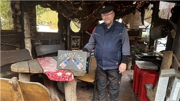 Ein Mann steht in einer rustikal eingerichteten Hütte und hält eine Hand hinter einem Schild, das auf einem Tisch steht. Auf dem Schild steht "Stammtisch" geschrieben, darunter sind zwei Glühweinflaschen zu sehen. Der Mann trägt ein gestreiftes Oberteil unter einer dunkelblauen Weste, ein schwarze Hose und eine schwarze Mütze.