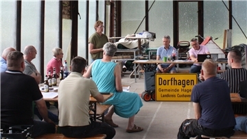 Einwohnerversammlung in Dorfhagen: Mehrere Personen sitzen an Tischen in einer Halle, eine Frau steht und spricht, im Vordergrund ein Ortsschild von Dorfhagen.