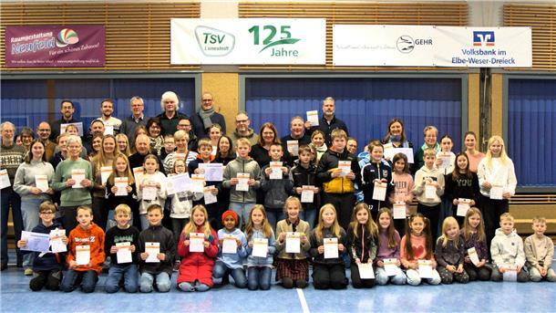 96 Sportabzeichen in Lunestedt verliehen - Gisela Hering führt die Rangliste an Kinder und Erwachsene knien und stehen in einer Sporthalle. In ihren Händen halten sie Urkunden.
