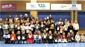 Kinder und Erwachsene knien und stehen in einer Sporthalle. In ihren Händen halten sie Urkunden.