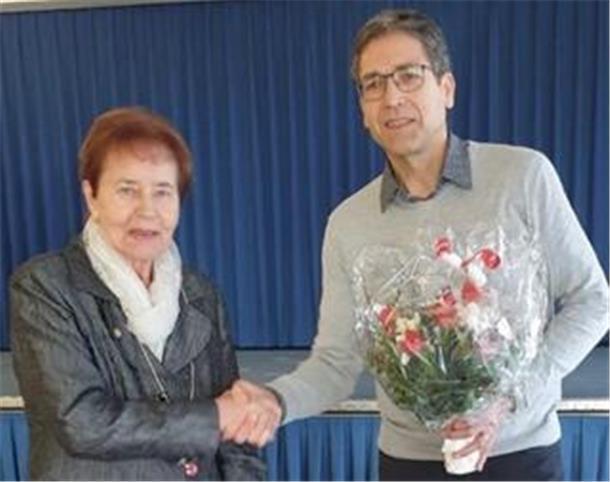 Ein Mann mit Brille, grauem Pullover und dunkler Hose überreicht einer älteren Frau mit kurzen rötlichen Haaren, schwarzer Jacke und weißem Schal einen Blumenstrauß. Beide schütteln sich die Hände.