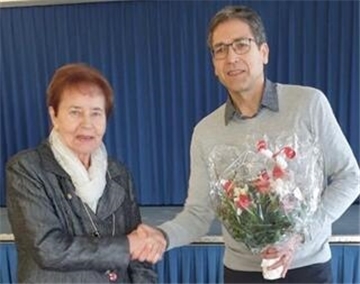 Ein Mann mit Brille, grauem Pullover und dunkler Hose überreicht einer älteren Frau mit kurzen rötlichen Haaren, schwarzer Jacke und weißem Schal einen Blumenstrauß. Beide schütteln sich die Hände.