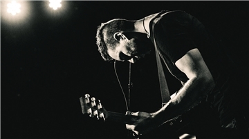 Musiker Troy Petty mit Gitarre auf einem Schwarz-Weiß-Foto im Scheinwerferlicht.