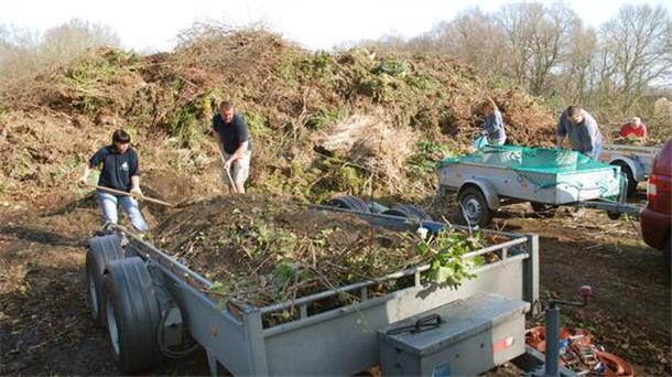 Mehrere Personen arbeiten im Freien auf einer Grünabfall-Deponie. Sie beladen oder entladen Anhänger mit Gartenabfällen wie Ästen, Laub und Erde.