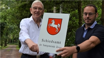 Zwei Männer halten ein Schild mit der Aufschrift Schiedsamt der Samtgemeinde Zeven.  