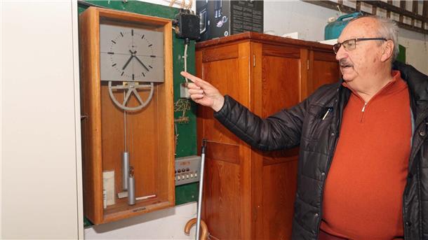 Ein Mann vor einer Uhr. Zu sehen sind auch ein Zahnrad und zwei kleine Gewichte.