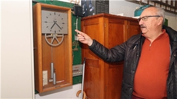 Ein Mann vor einer Uhr. Zu sehen sind auch ein Zahnrad und zwei kleine Gewichte.