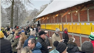 Menschen stehen an einem alten Gebäude und vergleichen an gelben Tafeln, ob sie bei der Lotterie gewonnen haben. 