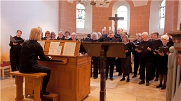 Eine Frau sitzt in einer Kirche an einer Orgel. Ihr steht ein Chor gegenüber und singt. 
