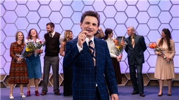 Hans Rosenthal (Florian Lukas) gestikuliert lächelnd im bunten "Dalli-Dalli"-Fernsehstudio. Hinter ihm stehen einige gut gelaunte Kandidatinnen und Kandidaten (Komparsen). Das biografische Drama „Rosenthal“ wird am Montag, 7. April (20.15 Uhr), im ZDF gezeigt.