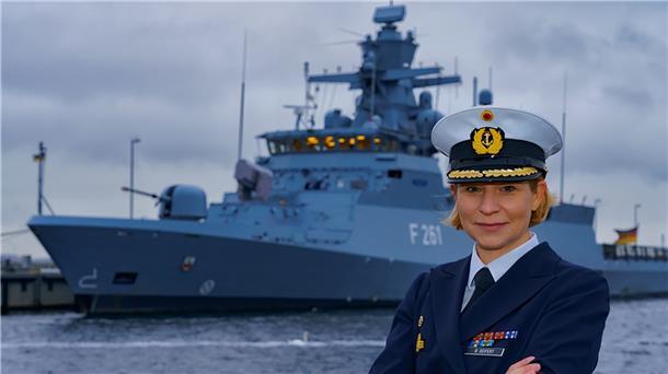 Bianca Seifert ist Deutschlands erste Korvettenkommandantin Bianca Seifert blickt in die Kamera. Im Hintergrund liegt eine Korvette vor Anker.