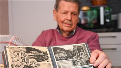 Ein Mann mit rostrotem Pullover, Oberlippenbart und grauen Haaren schaut in ein Fotoalbum. Er sitzt an einem Tisch.
