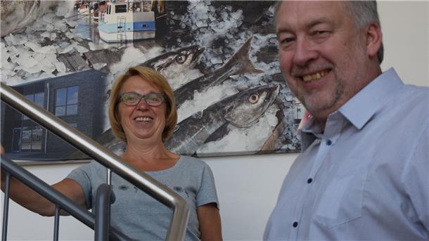 Ein Mann und eine Frau stehen auf einer Treppe. Im Hintergrund hängt ein Bild, auf dem Fische und Eis gemalt sind.