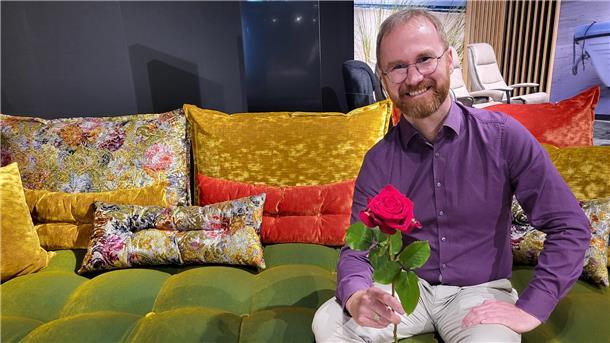 Ein Mann sitzt lächelnd auf einem bunten Sofa und hält eine Rose in der Hand. 