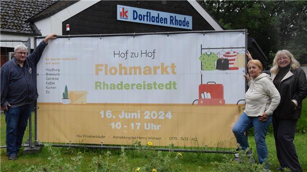 Henry Wichern vom Organisationsteam sowie Gisela Marquarding und Birgit Arndt (rechts) von den Rhader Landfrauen am Banner, das im Rhader Ortskern am Dorfladen für den Flohmarkt in Rhadereistedt wirbt.