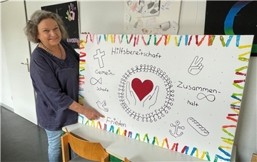 Eine Frau, die ein Plakat in die Kamera hält. Die Frau hat eine lilafarbene Bluse an. Auf dem Plakat ist ein rotes Herz aufgemalt, dazu die Begriffe Hilfsbereitschaft, Gemeinschaft, Zusammenhalt und Frieden.
