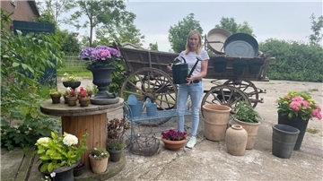 Frau mit Gießkanne vor einem alten Holzwagen, rundherum Blumentöpfe.