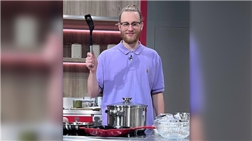 Ein junger Mann mit Brille und blondem Bart steht in einer modernen Küche. Er trägt ein lila Poloshirt mit einem kleinen Logo und hält eine schwarze Pfannenwender hoch. 