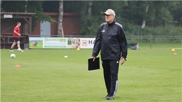 Ein Mann in einem schwarzen Sportdress und einer Cap hält eine Mappe in der Hand und geht über einen Rasen.