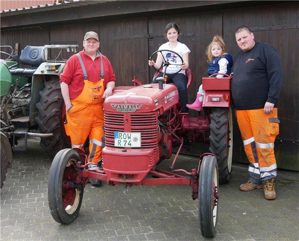 Zwei Kindern sitzen auf einem kleinen roten Traktor. Daneben stehen zwei Männer.