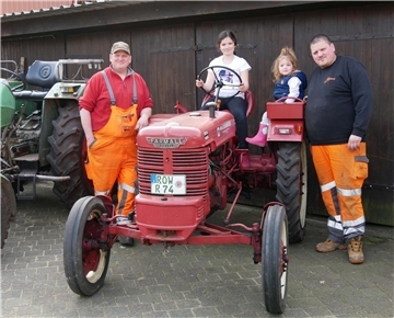 Zwei Kindern sitzen auf einem kleinen roten Traktor. Daneben stehen zwei Männer.
