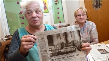 Ein Mann hält eine Zeitungsseite in die Kamera. Daneben sitzt eine Frau im Hintergrund.