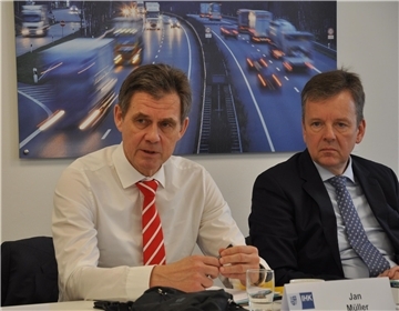 Zwei Männer sitzen an einem Tisch. Im Hintergrund hängt das Foto einer Autobahn.