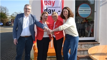 Ein Mann und drei Frauen stehen vor einem Plakat mit der Aufschrift "Equal Pay Day" und legen ihre Hände übereinander.