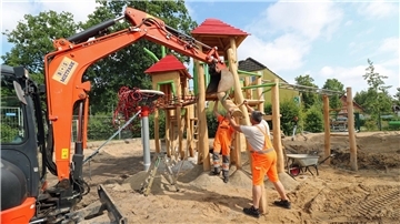 Zwei Bauarbeiter montieren mit Hilfe eines Baggers ein neues Holzspielgerät mit Rutsche auf einem sandigen Schulhofgelände. Im Hintergrund sind weitere Spielgerüste und ein Wohnhaus zu sehen.