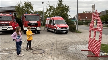 Zwei Kinder üben mit einem Feuerwehrschlauch das Zielspritzen auf ein stilisiertes Hausmodell, im Hintergrund stehen mehrere Feuerwehrfahrzeuge.