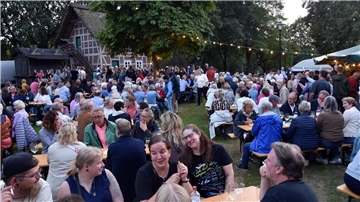 Unzählig viele Menschen sitzen an Biertischgarnituren vor einem norddeutschen Fachwerkhaus, im Hintergrund stehen auch viele Menschen.