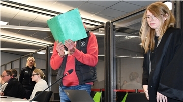 Mann mit Mappe vor dem Gesicht neben seiner Anwältin. 