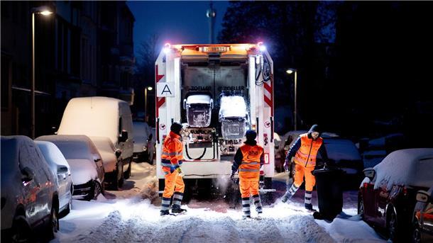 Mitarbeiter der Abfallwirtschaft leeren am frühen Morgen die Mülltonnen in einer völlig verschneiten Straße