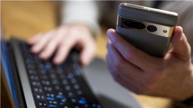 Auf dem Foto ist eine Hand zu sehen, die ein Smartphone hält, die andere liegt auf einer Computertastatur.