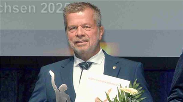Ulrich Seier im Anzug bei der Preisverleihung des „Großen Preises des Mittelstandes“. Er hält eine Urkunde, einen Blumenstrauß und eine silberfarbene Trophäe in der Hand.