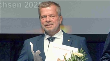 Ulrich Seier im Anzug bei der Preisverleihung des „Großen Preises des Mittelstandes“. Er hält eine Urkunde, einen Blumenstrauß und eine silberfarbene Trophäe in der Hand.